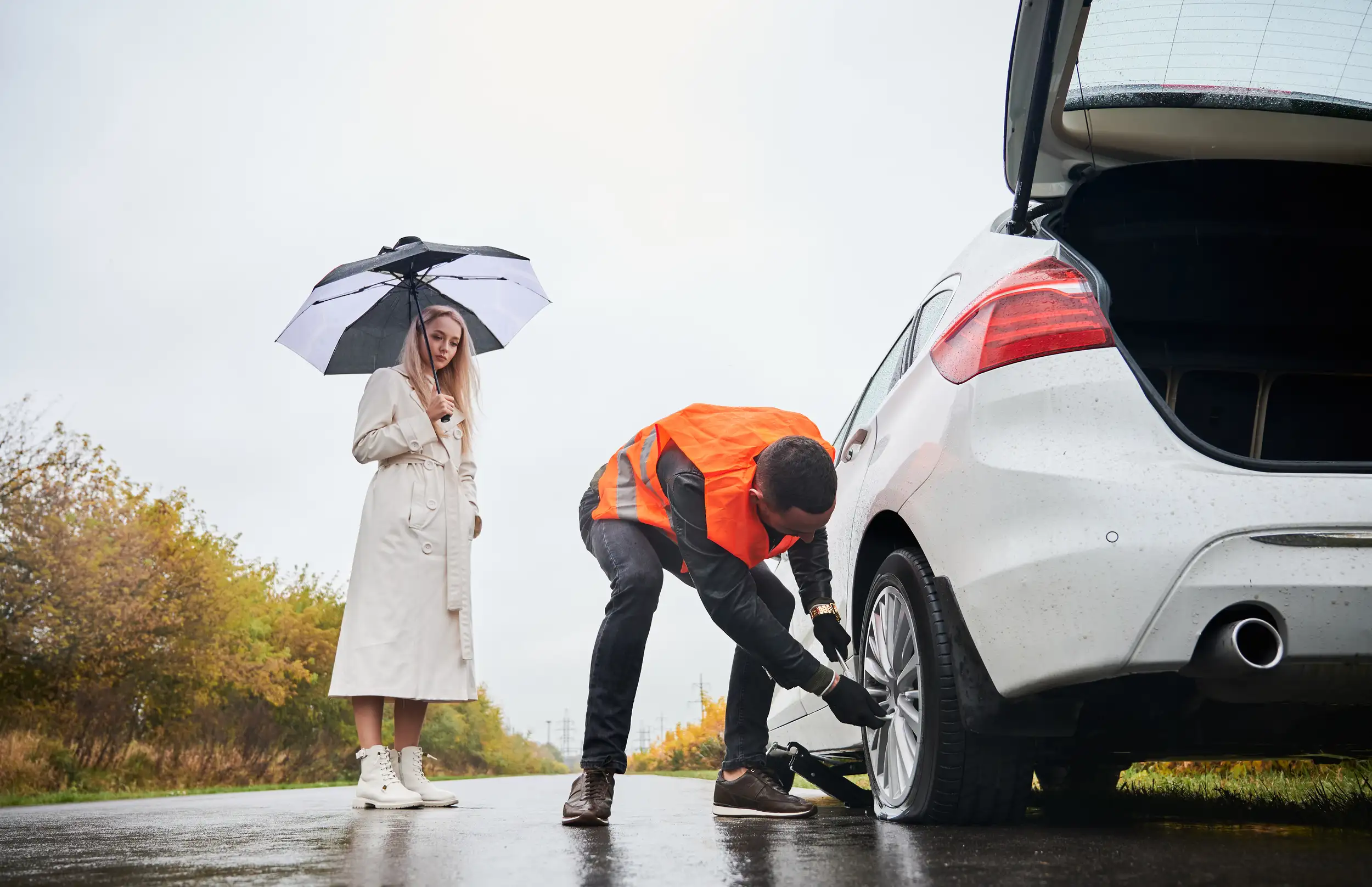 Roadside assistance worker in orange vest repairing flat tire for a woman holding an umbrella by her white vehicle on a rainy day.